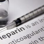 A syringe and a vial are placed on a document with the word Heparin prominently highlighted, along with explanatory text about the medication.