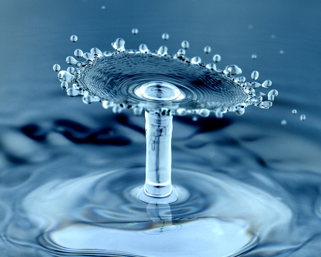 A close-up photo captures a water droplet collision, forming a mushroom-like splash with tiny droplets scattering around, set against a smooth blue water surface.